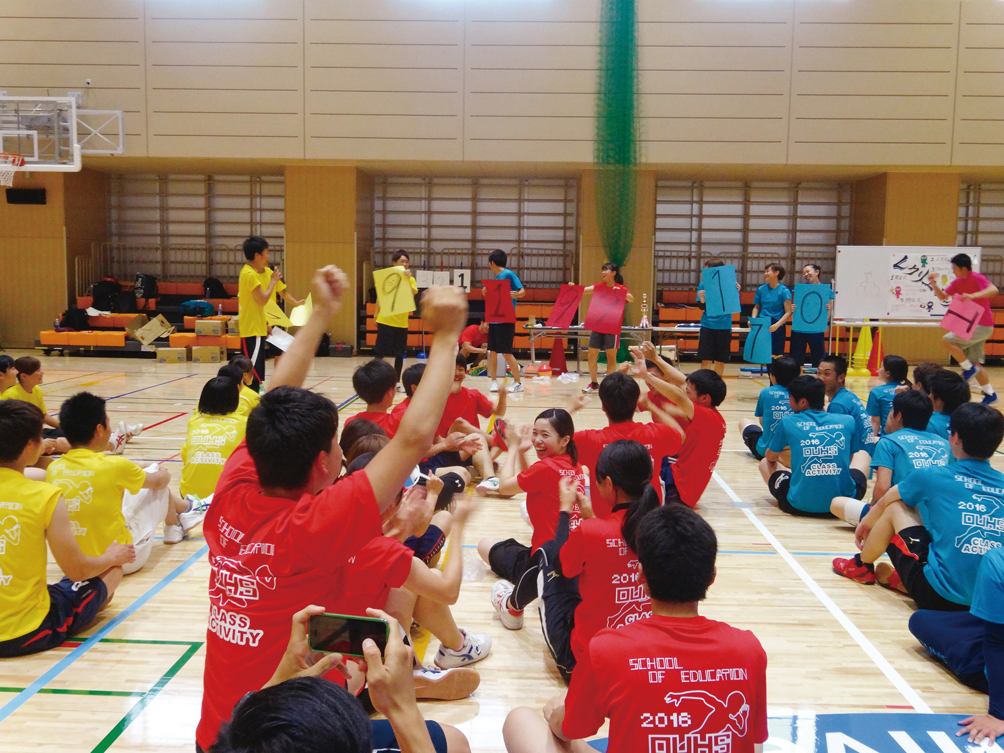 高齢者グループがとどろきアリーナでレクリエーションを楽しむ風景。自然光が差し込む体育館で、全員が輪になって手をつなぎ、インストラクターに合わせて運動している。